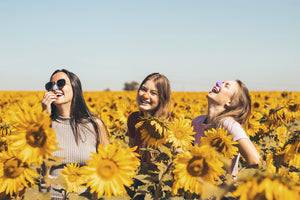 Three girls wearing the best Noz reef safe sunscreen for face in the colors blue, purple, and orange laughing in a sunflower field.