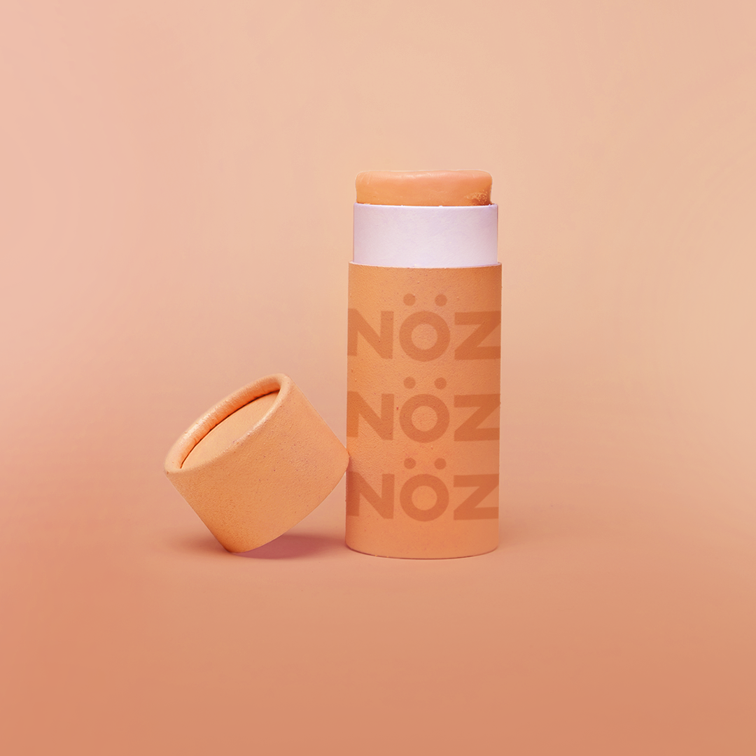 An open tube of orange Nöz sunscreen with its lid tilted beside it and shown against a orange background